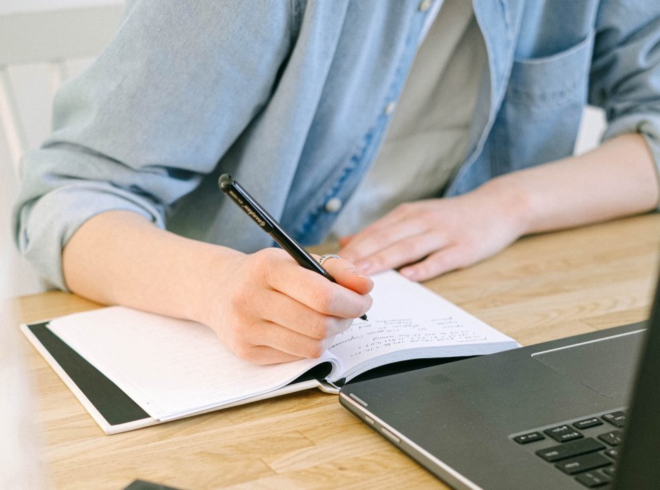 Person beim Schreiben in ein Notizbuch neben einem Laptop.
