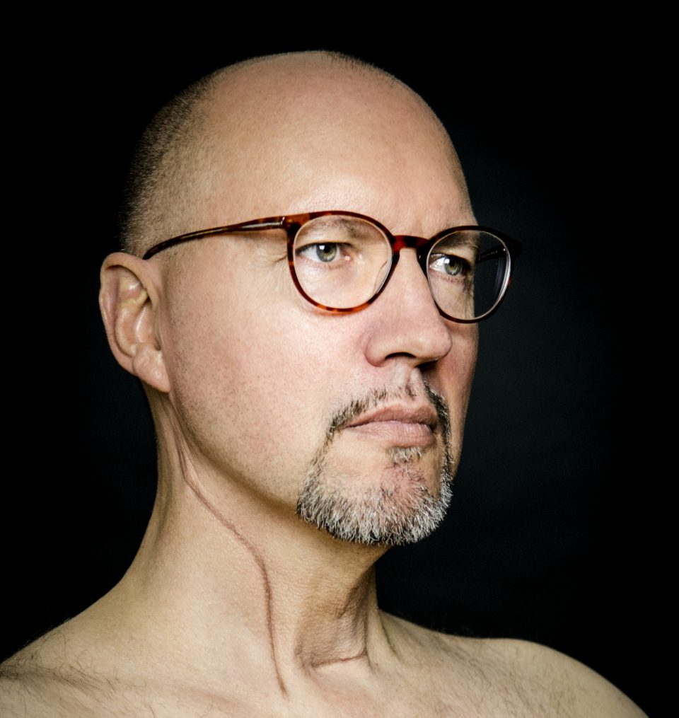Mann mit Glatze und Brille vor schwarzem Hintergrund.