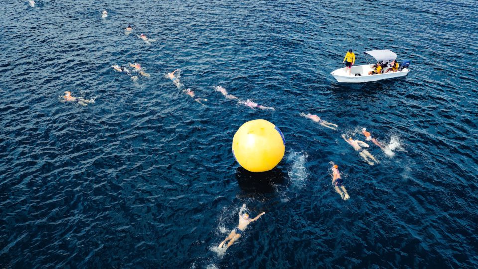 Mehrere Menschen schwimmen um eine gelbe Boje in Form eines großen Ballons, beobachtet von einem Wettrichter auf einem Boot stehend.
