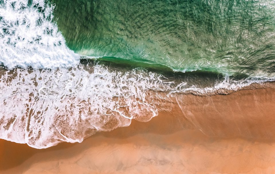 Ein Strand am Meer von oben, grün schimmerndes Wasser geht in weiß schäumende Wellen über, die über den dunkel- und hellbraunen Sand fließen.