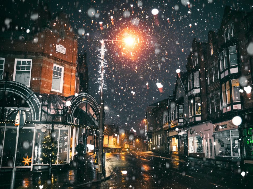 Nächtliche Häuserzeilen einer Kleinstadt bei Schneefall, von einzelnen Straßenlaternen besinnlich beleuchtet.