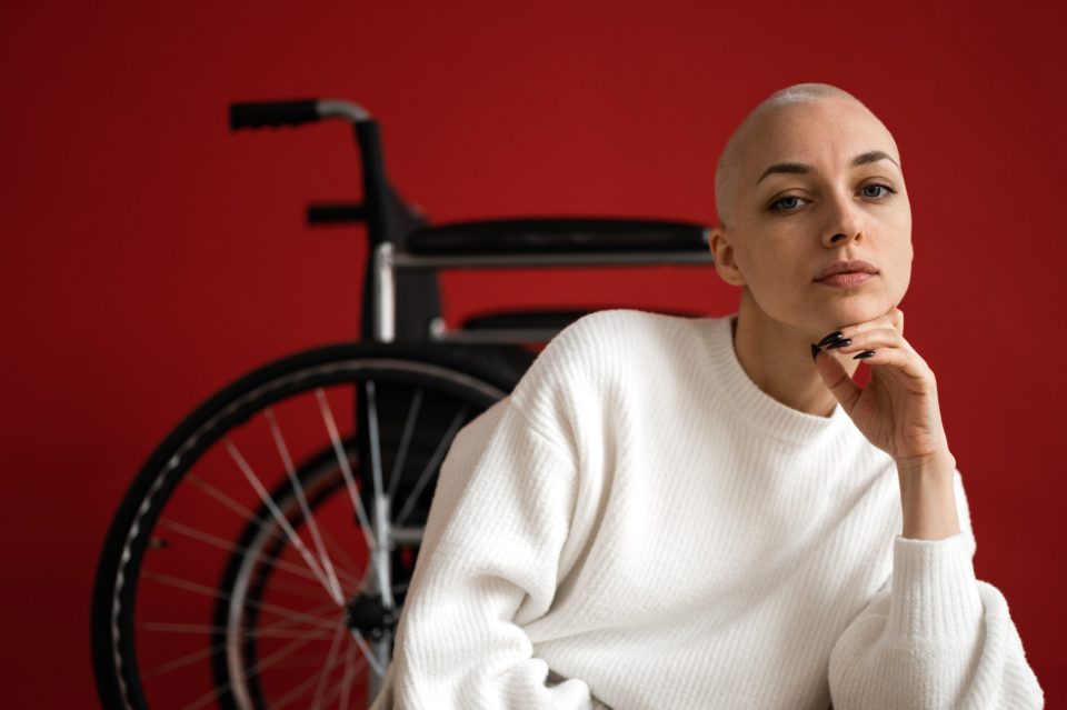 Junge Frau in weißem Pullover sitzt in selbstbewusster Pose vor einem Rollstuhl, dahinter eine rote Wand.