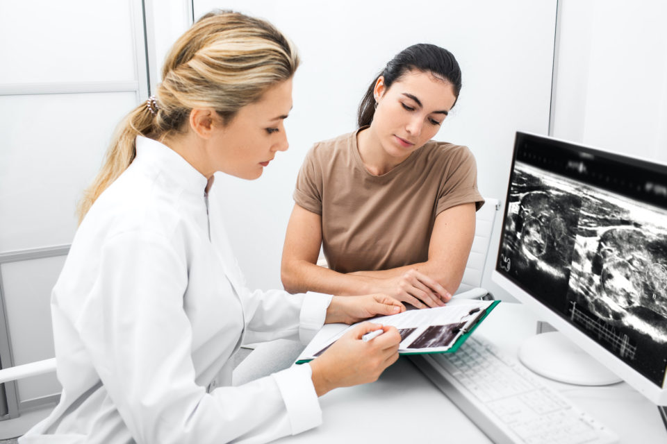 Eine junge Ärztin erklärt einer jungen Patientin Ultraschallbilder auf einem Ausdruck vor einem Monitor.