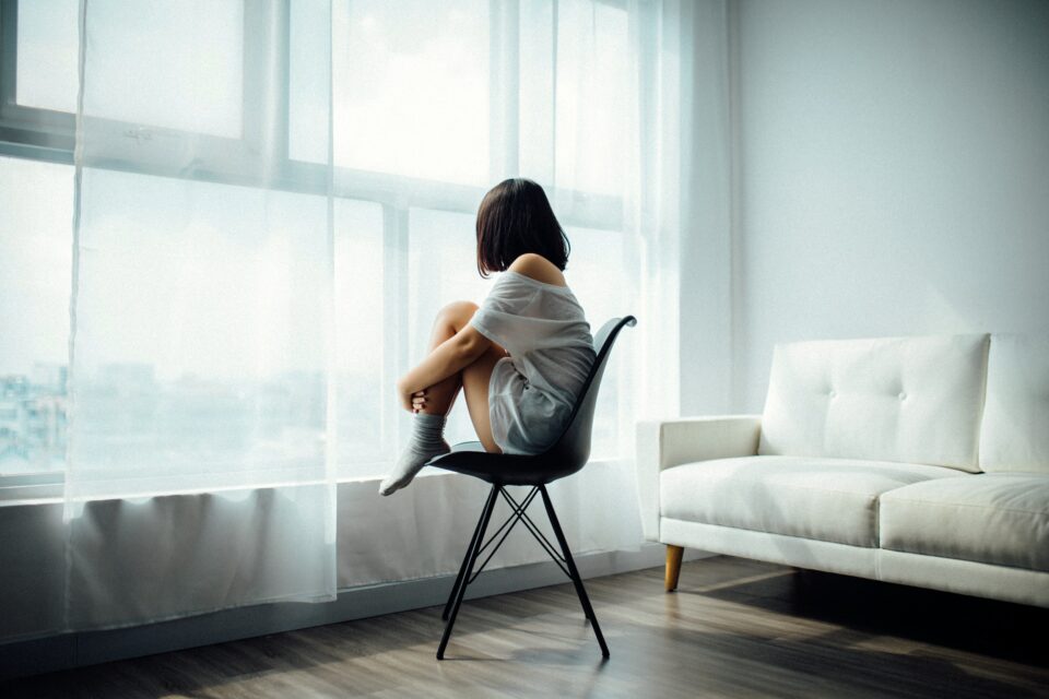 Frau in zusammengekauerter Haltung auf einem Stuhl, blickt aus dem Fenster.