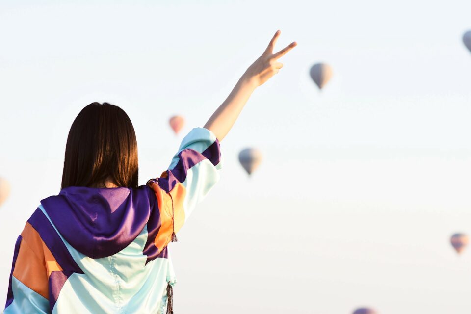 Frau streckt den Arm einigen entfernten Heißluftballons entgegen, formt mit den Fingern das Vivat-Zeichen.