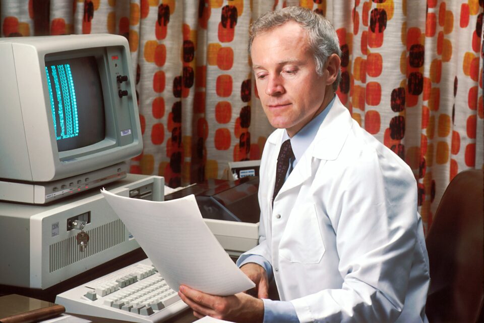 Arzt, der vor einem veralteten Computer sitzt und auf Dokumente starrt