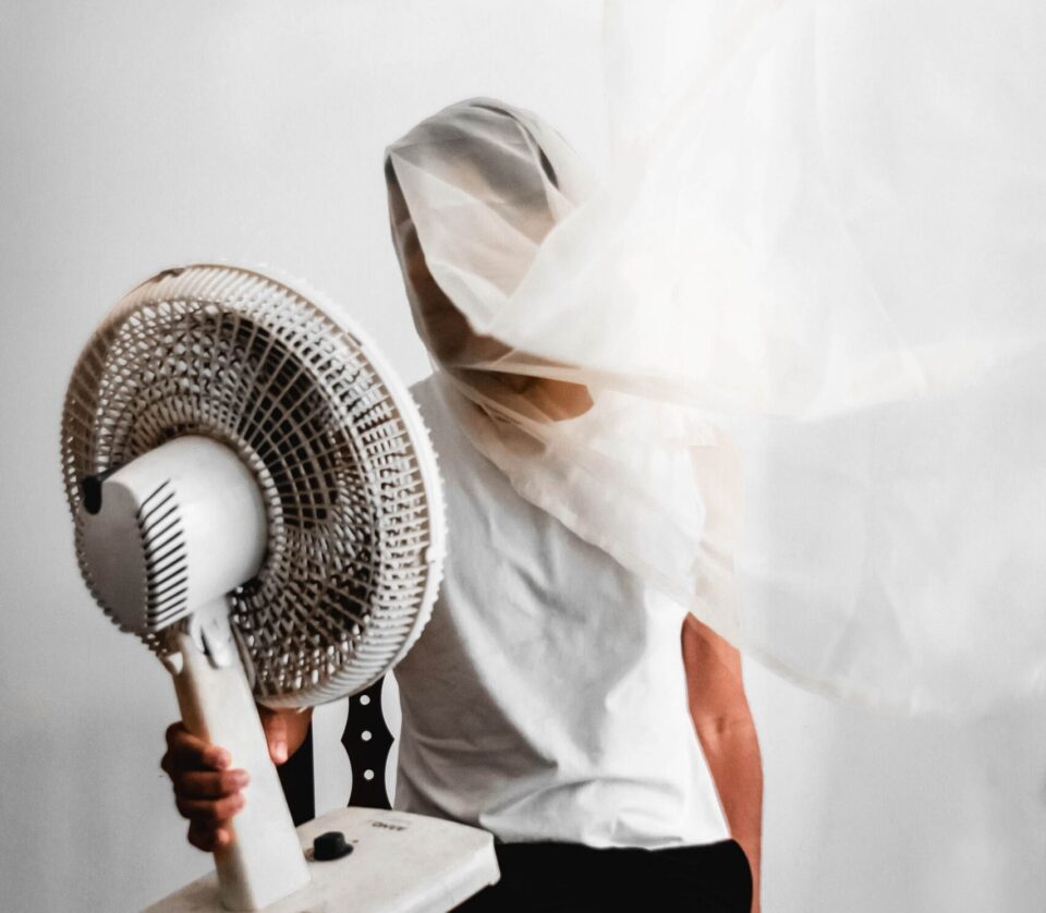Person mit leichtem weißen Tuch vor Gesicht hält Ventilator