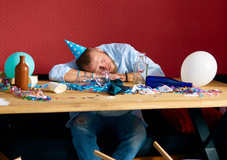 Man Sleeping At Table With Blue Cap In Messy Room After Birthday Party